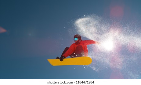 CLOSE UP: Snowboarder Jumping Big Air Kicker, Spraying Snowflakes And Flying Over Sun On Perfect Winter Day. Snowboard Jump In Snow Park. Sunbeams Shining Past Jumping Boarder In Mountains