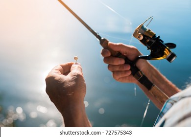 Close Up. Spinning With The Fishing Reel In The Right Hand, Fishing Hook On The Line With The Bait In The Left Hand Against The Background Of The Water, In Which The Sun Is Reflected