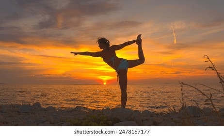 CLOSE UP: Strong girl balancing in king dancer position on stunning rocky beach by rippling sea under magical golden setting sky. Woman in standing yoga pose with backbend, holding leg with one hand - Powered by Shutterstock