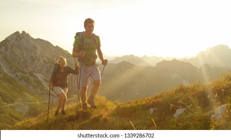 CLOSE UP, SUN FLARE: Happy Tourists Hiking In The Stunning Mountains Of Slovenia On A Sunny Summer Day. Athletic Couple Trekking In The Julian Alps At Sunrise. Man And Woman Exploring Golden Landscape