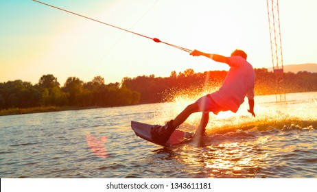 CLOSE UP, SUN FLARE: Unrecognizable athletic man wakesurfing on the lake at golden sunset. Young surfer dude having fun wakeboarding in the cable park on a beautiful summer morning. Fun water sport