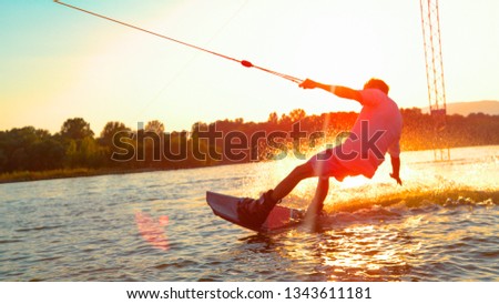 CLOSE UP, SUN FLARE: Unrecognizable athletic man wakesurfing on the lake at golden sunset. Young surfer dude having fun wakeboarding in the cable park on a beautiful summer morning. Fun water sport