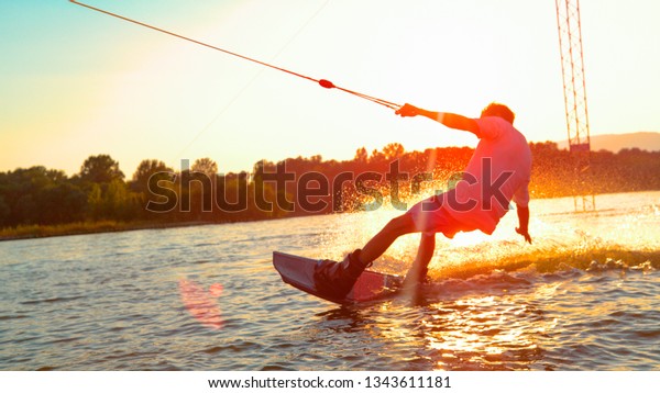 CLOSE UP, SUN FLARE: Unrecognizable athletic man
wakesurfing on the lake at golden sunset. Young surfer dude having
fun wakeboarding in the cable park on a beautiful summer morning.
Fun water sport
