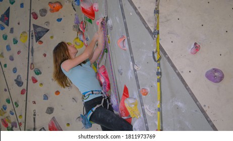 CLOSE UP: Teenage Girl Climbing Indoors Is Trying To Clip The Belay Rope In A Safety Carabiner. Experienced Caucasian Teenager Climbing A Difficult Route At An Indoor Rock-climbing Training Facility.