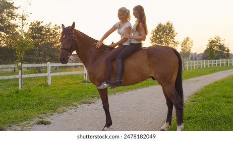 CLOSE UP: Two beautiful blonde Caucasian girls sitting on strong chestnut gelding at golden sunset. Happy girlfriends horseback riding stunning brown horse on ranch at amazing sunrise - Powered by Shutterstock