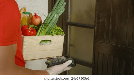 CLOSE UP: Unrecognizable young man wearing latex gloves holds a big wooden box of produce and a payment terminal while waiting for the door to open. Delivery guy in red is waiting by the closed door. - Powered by Shutterstock