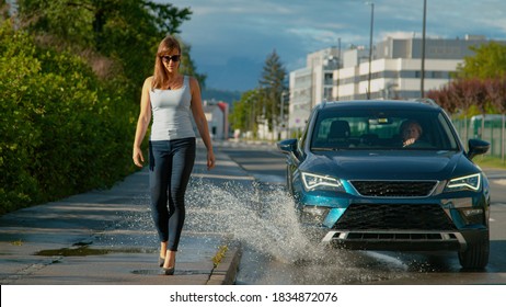 CLOSE UP: Unsuspecting Businesswoman Walking Along Sidewalk Gets Splashed By Careless Driver Driving Down The Empty Road. Senior Woman Drives Her Blue SUV Into A Puddle, Splashing Water At Pedestrian