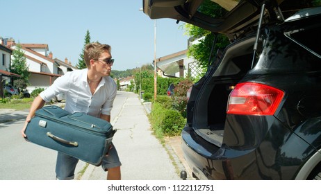 CLOSE UP: Whistling Young Male Tourist Throwing His Bags Into Big Black SUV Before Driving Away From Home To Go On Awesome Road Trip. Carefree Caucasian Man Packing For Summer Holiday With Friends.