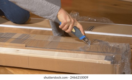 CLOSE UP: Woman cuts and peels away plastic foil off a piece of furniture shipped inside a cardboard box. Young female with red nails unboxes wooden pieces of furnishing delivered to her new home. - Powered by Shutterstock