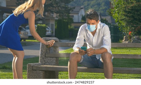 CLOSE UP: Young Caucasian woman scares away stranger by spraying him with sanitizer before she sits down on the public bench. female wearing a facemask sprays man sitting on the bench and texting. - Powered by Shutterstock