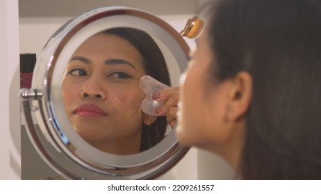 CLOSE UP: Young Lady Self Massaging Her Face With Jade Scraper For Facial Lifting. Reflection Of Beautiful Asian Woman In Cosmetic Mirror Performing Anti-aging Massage Treatment With Gua Sha Scraper.