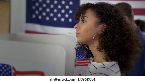 Close Up, Young Latina Woman Looking Thoughtful As She Considers Vote At Polling Station. Other Voters In Background And Large US Flag On Wall Behind