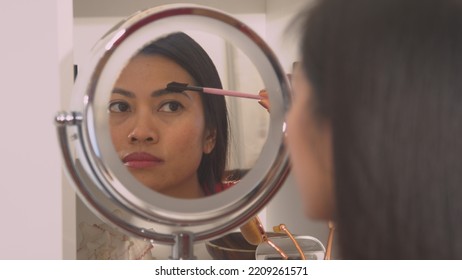 CLOSE UP: Young Woman Correcting Eyebrow Shape With Brush In Mirror Reflection. Pretty Philippine Lady Beautifying And Shaping Her Eyebrows With Brow Comb. Female Person Perfecting Her Face Make Up.