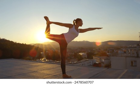 CLOSE UP: Young woman doing lord of the dance pose on left foot on the top of skyscraper standing on edge looking down on stunning green overgrown mountains and vivid town at sunset - Powered by Shutterstock