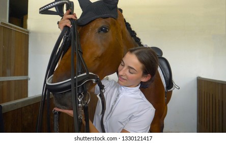 CLOSE UP: Young woman tacking up and putting a bridle on her beautiful brown horse before a dressage competition. Happy Caucasian girl sliding a bit into her stallion's mouth while tacking him up.