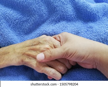 Close Up, Younger Hand Is Holding Elder Hand On Blue Blanket. Palliative Care Concept. 