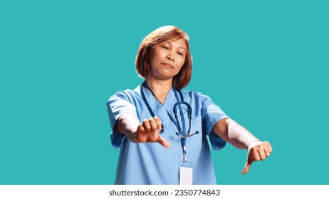 Close up of upset hospital worker showing thumbs down signs while at work, isolated over studio background. Dissatisfied asian healthcare expert doing frantic disapproval gestures
