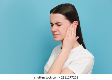 Close up of upset sad young millennial female holding painful ear, suddenly feeling strong ache, posing isolated on blue color background studio with copy space for advisement. Health problem concept