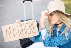 Close Up Upset, Tired, Sad Blond Woman Pulling Hat Over Eyes, Holding Carton Board Sign Anywhere With Luggage Hitchhiking, Sitting By The Road. Adventure Alone, Autostop And Hitchhiking Lifestyle
