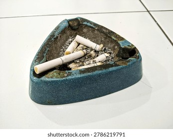 Close up of used blue ceramic triangular ashtray filled with cigarette butts and ash on white tiled floor