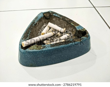 Close up of used blue ceramic triangular ashtray filled with cigarette butts and ash on white tiled floor