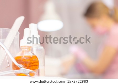 Close up of various professional cosmetics and oils, tools for pedicure on a white table. View from a distance of a specialist wearing gloves pedicuring for a client in an armchair. Beauty saloon