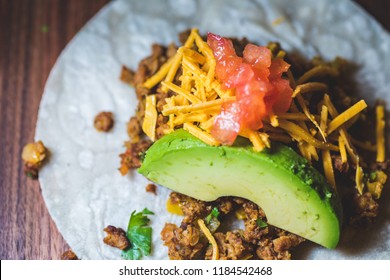 A Close Up Of A Vegan Taco With Ground Meat, Avocado, Cheese, And Diced Tomato On A Wood Board.