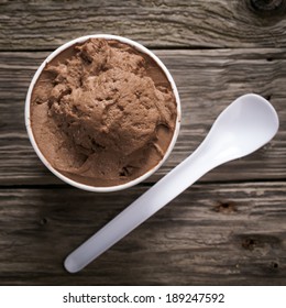 Close Up View From Above Of A Tub Of Creamy Italian Chocolate Or Mocha Ice Cream Served With A Disposable Plastic Spoon On An Old Textured Wooden Table, Square Format