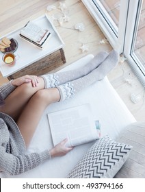 Close Up View From Above Of Woman Sitting Home By The Window On A Living Room Sofa Reading Magazine. Christmas Lights On The Floor, Books And Sweets On The Table