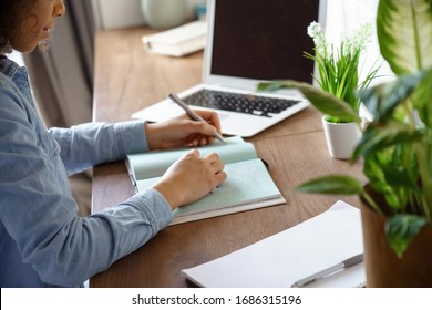 Close up view of african american teen girl student writes notes in diary notebook makes goals check list, agenda plan, distance studying concept with laptop mock up screen sits at home office desk.