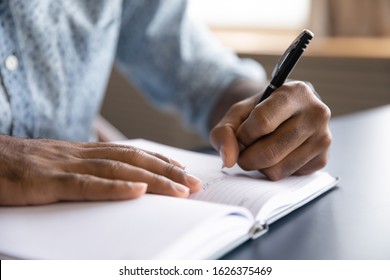 Close up view of african left-handed businessman writing in notebook, american male hands holding pen making notes planning new appointments information in organizer personal paper planner at desk