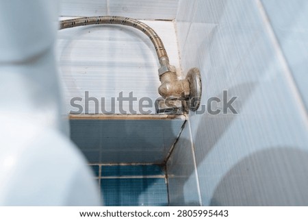 Close view of aged angled water shutoff valve connected to braided supply hose in tiled bathroom corner, showing mineral deposits, grout lines, rust stains, corrosion, and narrow maintenance access