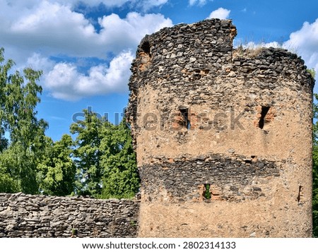 Close view of aged circular stone tower ruin showing layered stonework, cracked mortar, narrow arrow slit openings, and grass growing from upper crevices beneath bright midday sky with scattered