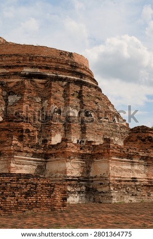 Close up view of the ancient red brick base of a stupa ruin at Wat Kudi Dao in Ayutthaya, Thailand. The historic architecture features weathered white stucco details under a bright cloudy blue sky