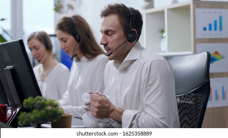 Close Up View Of Angry And Nervous Call Center Operator With Headset Talking With The Hotline Customer, Being Irritated, Uses Bubble Wrap To Calm Down And Continues Conversation. Stressful Situation