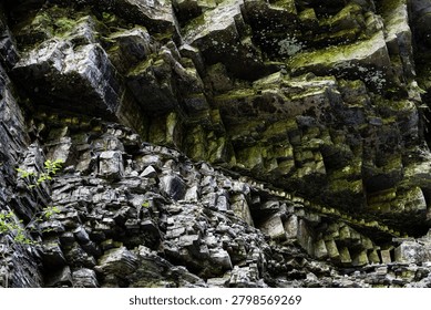Close view of angular rock shelves and cavities in a shaded cliffside reveals striking textures and mossy growth forming a rugged natural pattern shaped by water and time