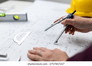 Close view of an architect using a compass to mark dimensions on a printed building plan near a yellow safety helmet.