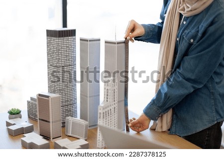 Close up view of architectural models presenting innovative metropolitan area set on office desk indoors. Female architect in casual clothes using metal ruler for measuring multi-storey building.
