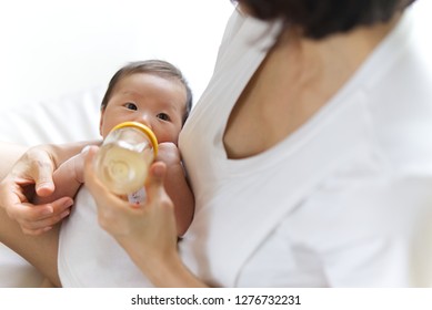 Close Up View Of Asian Mother Holding And Feeding Baby From Milk Bottle. Portrait Of Cute Newborn Baby Being Fed By Her Mother Using Bottle On Sofa. Loving Woman Giving To Drink Milk To Her Baby.