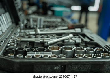 A close view of assorted socket wrenches, ratchets, and metal hand tools arranged in an open toolbox on a workbench, with a blurred industrial workshop in the