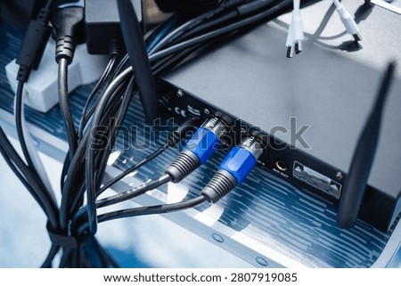 Close view of audio interface rear panel with blue XLR connector cables, tangled black wiring, and metal equipment case surface, lit by cool daylight for studio and broadcast use, with audio