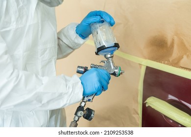 Close Up View Of Auto Painter In A Protective Suit And Mask Sprays A Degreaser Or Other Cleaner On A Car Door Using A Spray Gun. Preparation Of Vehicle Body Elements For Local Repair