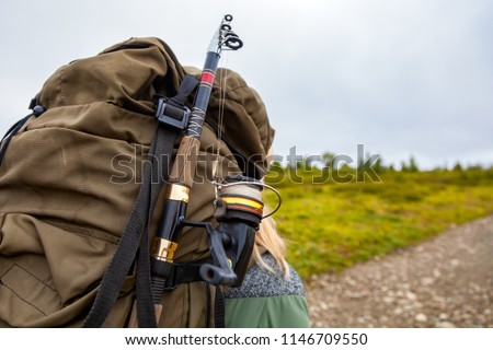 Close up view of back tourist  woman brown backpack  and fishing rod wearing on green tourism jacket  and walking along the path to the mountains on a sunny summer day