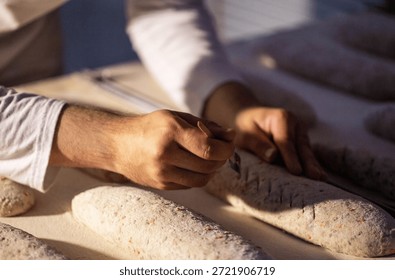 Close up view of baker hands manually scoring raw dough for bread production inside factory. Preparation of artisanal baguette loaves on bakery counter with flour.