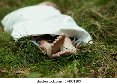 Close Up View Of Bare Feet Of Dead Body Wrapped In White Paper Or Film Laying On Green Grass