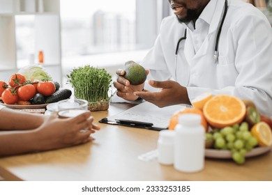 Close up view of bearded man holding green fruit while chatting with multiethnic person in office interior. Male nutritionist describing benefits of slimming foods during appointment in modern clinic. - Shutterstock ID 2333253185