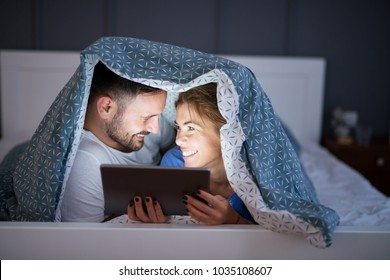 Nahaufnahme der schönen lächelnden jungen Frau und Ehemann, die nachts eine Tablette unter einer Decke auf dem Bett sehen. Beziehung und Ziele am Wochenende.
