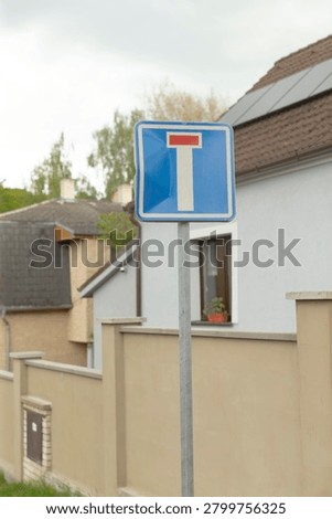 A close view of a blue dead end traffic sign mounted on a metal pole in front of suburban houses with plastered walls, tiled roofs and a small window with a