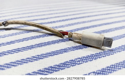 Close up view of a broken damaged USB Type-C charging cable exposing inner wires. This image shows the end of a severely damaged and frayed USB Type-C charging cable. Taken from a close-up, low angle, the photo reveals the cable's outer casing torn open to expose the internal red and black copper wires—which appear dull and worn—resting on a blue-and-white striped fabric.