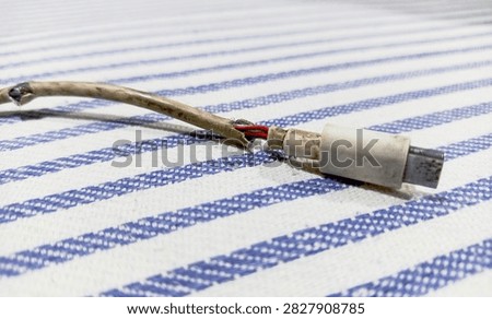 Close up view of a broken damaged USB Type-C charging cable exposing inner wires. This image shows the end of a severely damaged and frayed USB Type-C charging cable. Taken from a close-up, low angle, the photo reveals the cable's outer casing torn open to expose the internal red and black copper wires—which appear dull and worn—resting on a blue-and-white striped fabric.